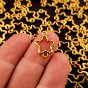 May include: Gold-toned metal star-shaped pendant with two jump rings. The pendant is held in a hand against a background of many similar star-shaped pendants.
