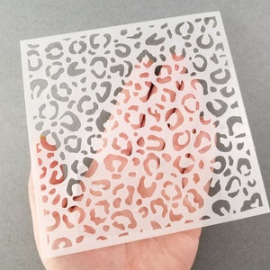 May include: A square, white stencil featuring a leopard print pattern. The stencil is held in a hand, and the design consists of irregular, rounded shapes. The background is a solid gray color.