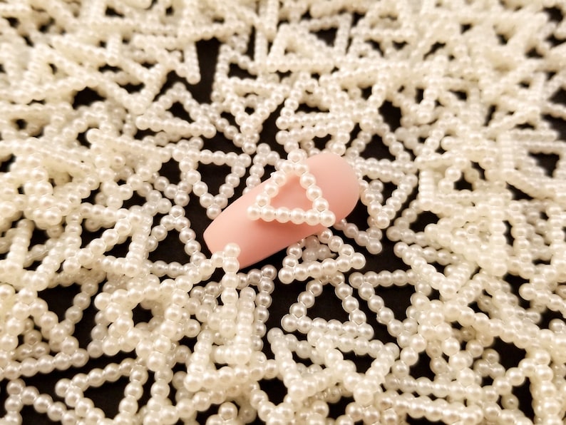 May include: A close-up of a pink nail with a white pearl triangle design. The nail is surrounded by a scattering of white pearl triangles.