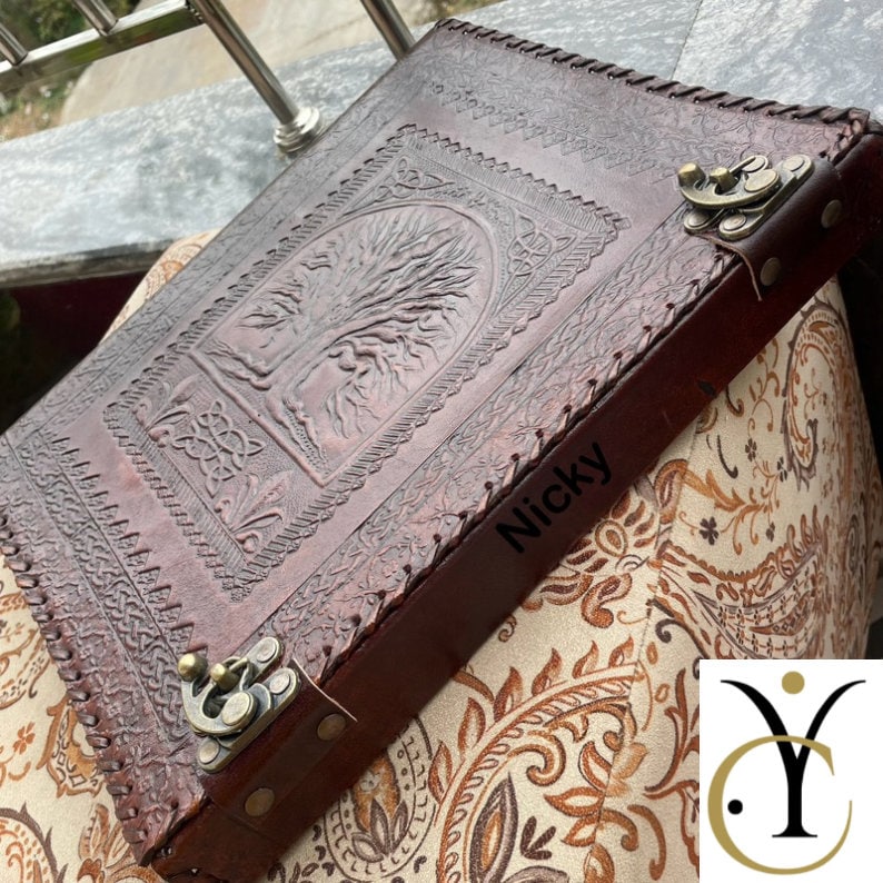 May include: A dark brown leather journal featuring an embossed tree design on the cover, intricate border patterns, and two antique brass-coloured metal latches. The name Nicky is printed on the side of the cover, which has stitched edges.