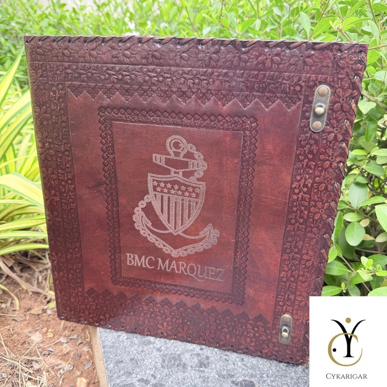 May include: A dark brown leather binder featuring an embossed floral border and a central engraved emblem of a shield and anchor. The text BMC MARQUEZ is etched below the emblem. The binder includes two metallic fasteners on the right edge.