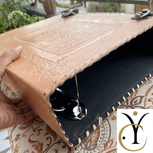 May include: A light tan leather binder featuring an embossed tree design on the cover, secured with two antique-style metal latches. The edges are stitched with leather lacing, and the interior is lined with black fabric, revealing a metal ring mechanism.