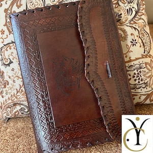 May include: A brown leather journal with intricate embossed designs and a lion's head. The journal has a flap closure and stitched edges. The cover features a detailed border and a central lion motif.