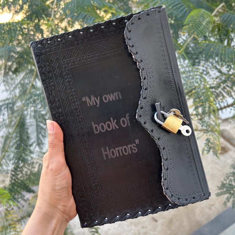 May include: Black leather journal with embossed floral patterns and the text "My own book of Horrors." The journal has a flap closure secured by a gold padlock and keys. The edges are stitched.