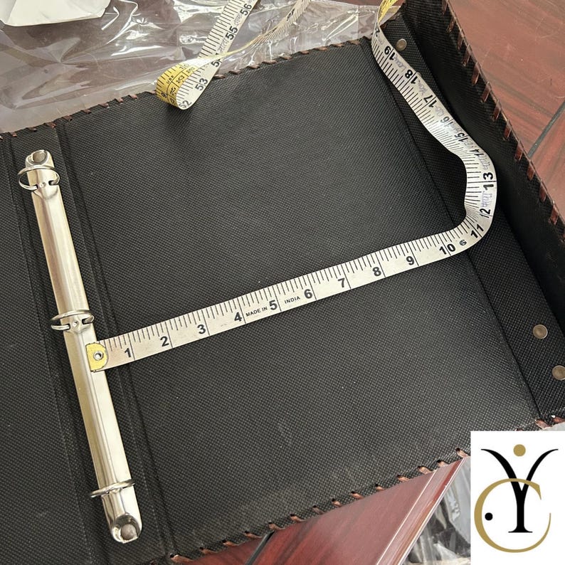 May include: A black three-ring binder with a textured surface and stitched edges. A white measuring tape is placed across the interior, showing a width of approximately 28 cm from the metal ring mechanism to the edge of the binder cover.