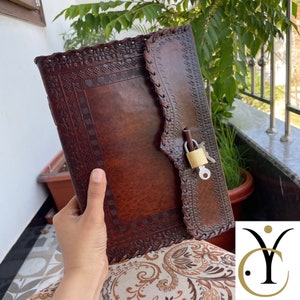 May include: A brown leather journal with intricate embossed designs and a decorative border. The journal is secured with a small gold padlock and key. The journal is held in a person's hand.