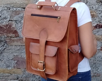 Può includere: Uno zaino in pelle marrone con una grande tasca frontale e una tasca più piccola sul lato. Lo zaino ha una chiusura a fibbia e una tasca con cerniera sul lembo superiore.