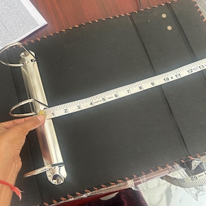 May include: A black three-ring binder with a textured surface and stitched edges. A white measuring tape is positioned across the metal ring mechanism, showing a width of approximately 31.75 cm from the spine to the edge of the binder cover.