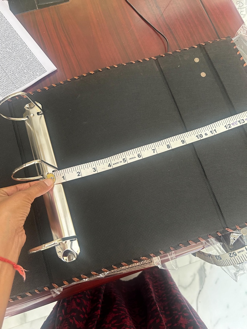 May include: A black three-ring binder with a textured surface and stitched edges. A white measuring tape is positioned across the metal ring mechanism, showing a width of approximately 31.75 cm from the spine to the edge of the binder cover.