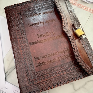 May include: A brown leather journal with a decorative border and a lock. The journal has engraved text that reads "Scott Allen Joseph Saved by Jesus" and "Noelle's Moments Photography". The journal is closed with a strap and a gold-colored padlock.