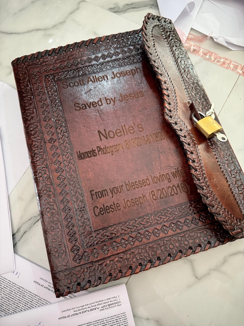 May include: A brown leather journal with a decorative border and a lock. The journal has engraved text that reads "Scott Allen Joseph Saved by Jesus" and "Noelle's Moments Photography". The journal is closed with a strap and a gold-colored padlock.