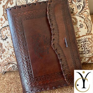 May include: A brown leather journal with intricate embossed designs and a lion's head. The journal has a flap closure and stitched edges. The cover features a detailed border and a central lion motif.