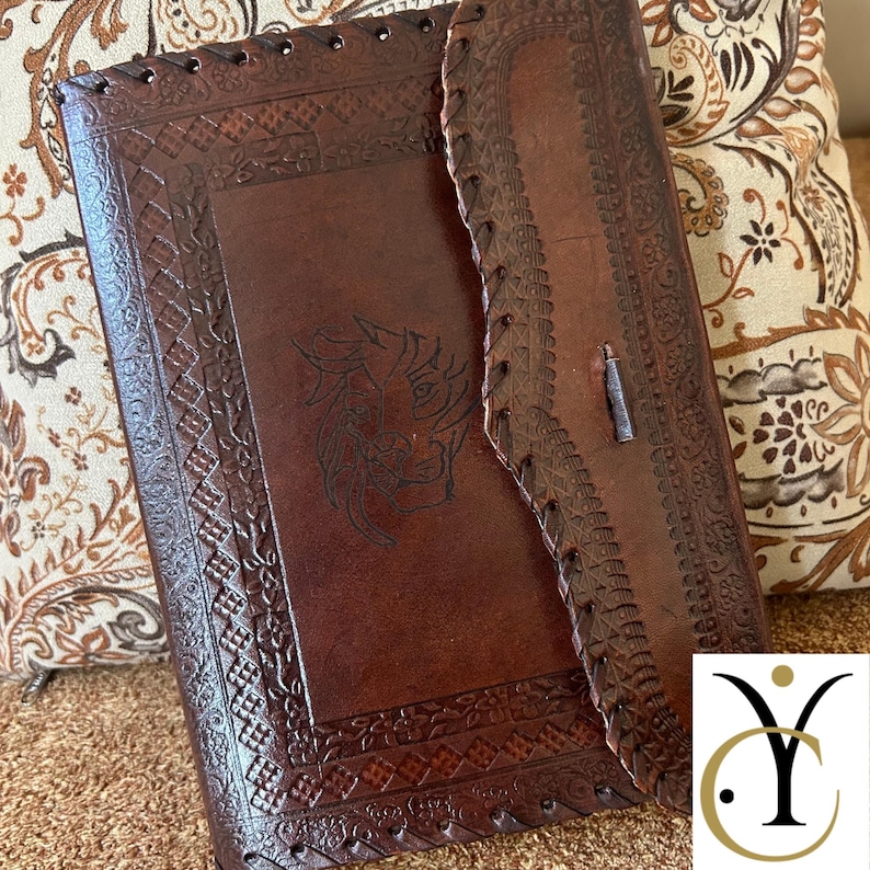 May include: A brown leather journal with intricate embossed designs and a lion's head. The journal has a flap closure and stitched edges. The cover features a detailed border and a central lion motif.