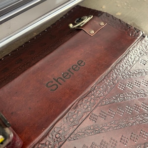 May include: A dark brown leather clipboard featuring a smooth central panel engraved with the name Sheree. The clipboard has a metallic clip mechanism at the top and decorative embossed geometric patterns along the side borders of the leather.