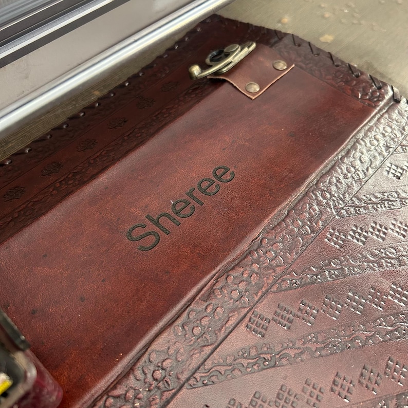 May include: A dark brown leather clipboard featuring a smooth central panel engraved with the name Sheree. The clipboard has a metallic clip mechanism at the top and decorative embossed geometric patterns along the side borders of the leather.