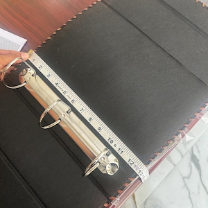 May include: A black three-ring binder with a textured cover and brown stitched edges. A silver metal ring mechanism is attached to the spine. A white measuring tape is placed across the mechanism, showing a length of approximately 31.75 centimetres.