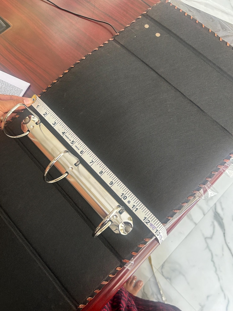 May include: A black three-ring binder with a textured cover and brown stitched edges. A silver metal ring mechanism is attached to the spine. A white measuring tape is placed across the mechanism, showing a length of approximately 31.75 centimetres.