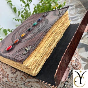 May include: A dark brown leather-bound journal featuring an embossed circular pattern on the cover, decorated with seven colorful oval stones arranged in a vertical line. The book has thick, deckled-edge cream pages and a metal latch closure.
