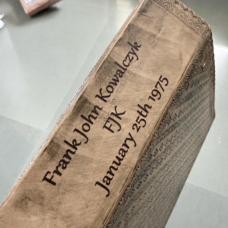 May include: A light brown wooden box with an engraved top surface featuring the text Frank John Kowalczyk, FJK, and January 25th 1975. The edges of the box are decorated with a carved, repeating geometric pattern along the entire perimeter.
