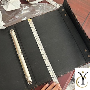 May include: A black three-ring binder with a textured surface and a silver metal mechanism. A white measuring tape is placed vertically across the centre, showing a length of 30.5 cm. The edges feature a decorative stitched pattern.