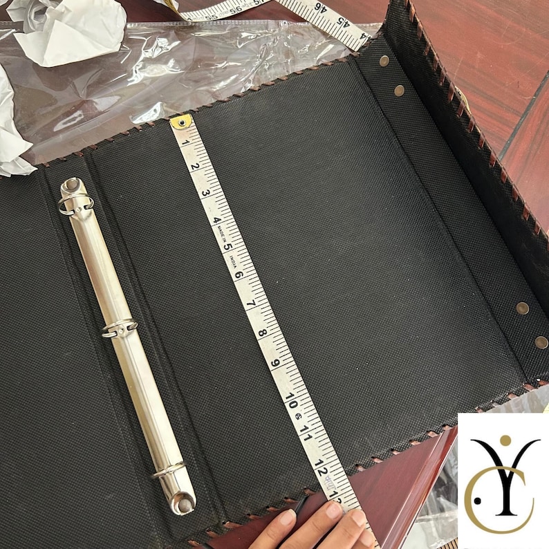 May include: A black three-ring binder with a textured surface and a silver metal mechanism. A white measuring tape is placed vertically across the centre, showing a length of 30.5 cm. The edges feature a decorative stitched pattern.