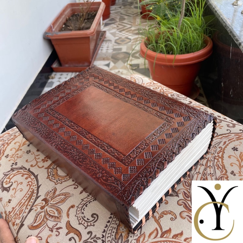 May include: A brown leather journal with intricate embossed designs on the cover. The journal has a rectangular shape with a thick stack of white pages. The cover features a central panel and decorative borders.
