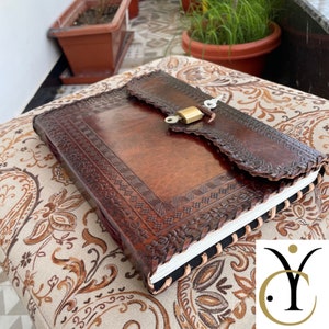 May include: A brown leather journal with a decorative embossed design and a flap closure secured by a brass lock. The journal's edges are hand-stitched, and the pages are visible. The journal is resting on a patterned cushion.