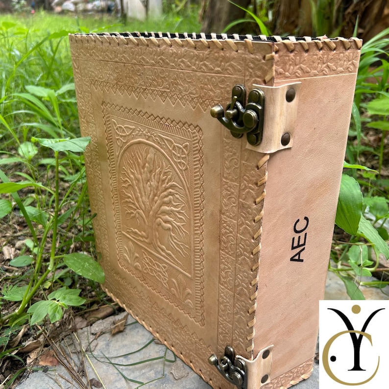 May include: A light tan leather-bound book featuring an embossed tree design on the front cover, intricate border detailing, and a metal latch closure. The spine of the book is clearly marked with the black capital letters AEC.