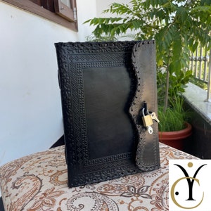 May include: Black leather journal with intricate floral and geometric embossed designs. The journal features a flap closure secured by a gold-colored padlock and key. The journal is resting on a patterned cushion.