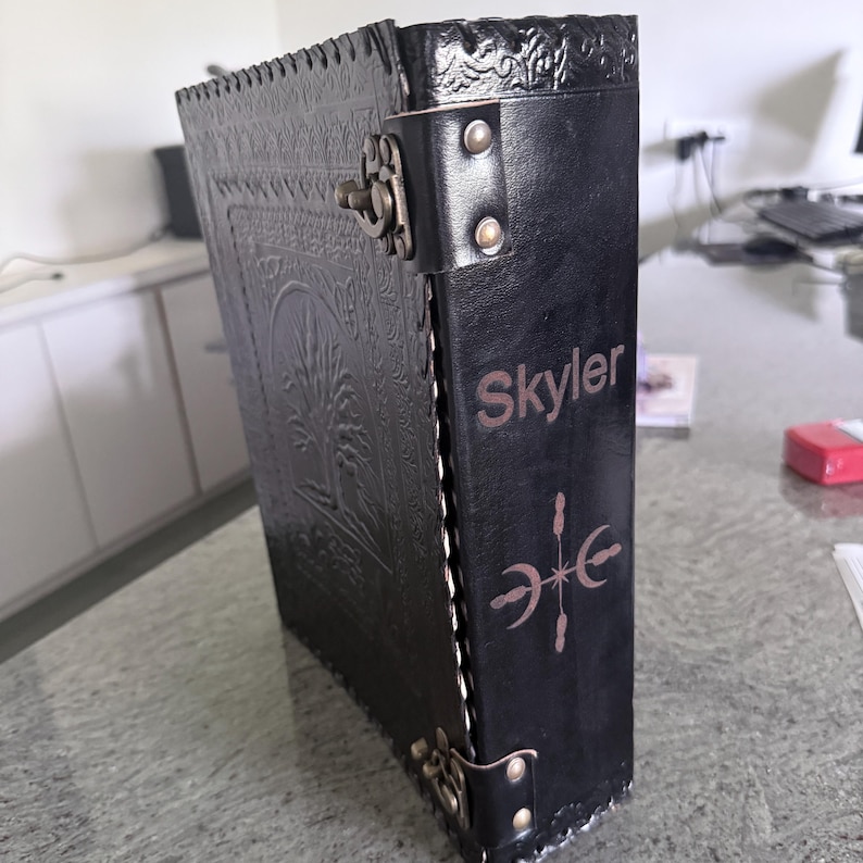 May include: A black leather-bound journal featuring an embossed tree design on the cover, a metal latch closure, and the name Skyler engraved on the spine above a celestial symbol. The book has decorative stitching along the edges and metal rivets.