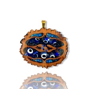 Könnte beinhalten: Ein Holzanhänger mit einer goldenen Öse und blauen bösen Augen-Anhängern, die in Harz eingebettet sind. Der Anhänger hat die Form einer halben Walnussschale und einen glitzernden blauen Hintergrund.
