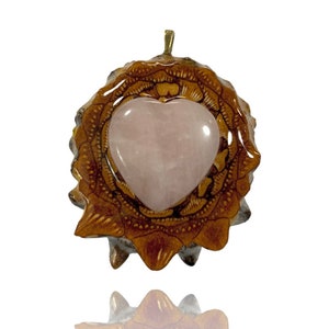 May include: A heart-shaped rose quartz pendant set in a brown wooden frame with intricate carvings. The pendant has a gold-colored bail.