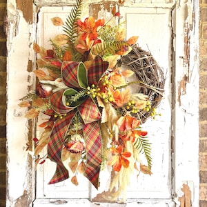 May include: A decorative wreath featuring a natural grapevine base adorned with artificial autumn foliage, including orange and yellow leaves, green ferns, wheat stalks, and small yellow berries, finished with a large plaid ribbon bow.