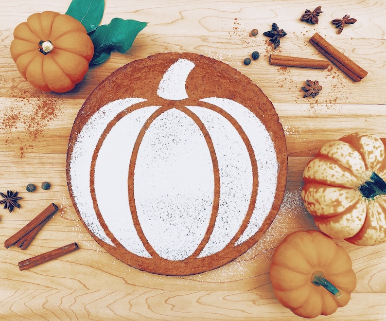 May include: A round pumpkin-shaped cake decorated with a white powdered sugar pumpkin design. The cake is surrounded by cinnamon sticks, star anise, and whole pumpkins.