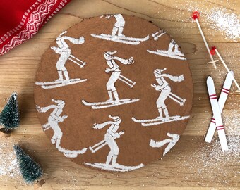 May include: A round chocolate cake decorated with white icing and skiers. The cake is on a wooden surface with two pairs of white skis and red ski poles.