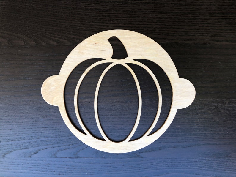 May include: A wooden pumpkin-shaped stencil with a circular cutout. The stencil is made of light-colored wood and has a simple design.