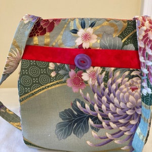 May include: A floral patterned shoulder bag with a red band and purple button closure. The bag features a mix of green, gold, and purple floral designs, with a light blue strap.
