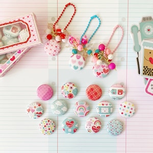 May include: A collection of Valentine's Day themed buttons and keychains. The buttons feature various designs, including hearts, flowers, and clouds. The keychains have colorful beads and charms. A decorative box with heart-shaped designs is also present.