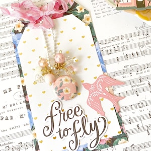 May include: A pink and white bird with a gold heart design flies above the words "Free to Fly" in black script. The background is a collage of floral patterned paper and sheet music.