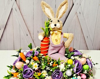 May include: A purple and green Easter centerpiece with a plush bunny holding a carrot. The centerpiece features purple roses, green foliage, and pastel colored eggs.