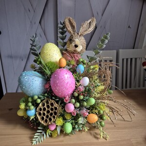 May include: An Easter centrepiece with a straw bunny, colourful eggs, and greenery. The arrangement includes large speckled eggs in blue, yellow, and pink, surrounded by smaller eggs and decorative elements. The centrepiece is set on a wooden table.
