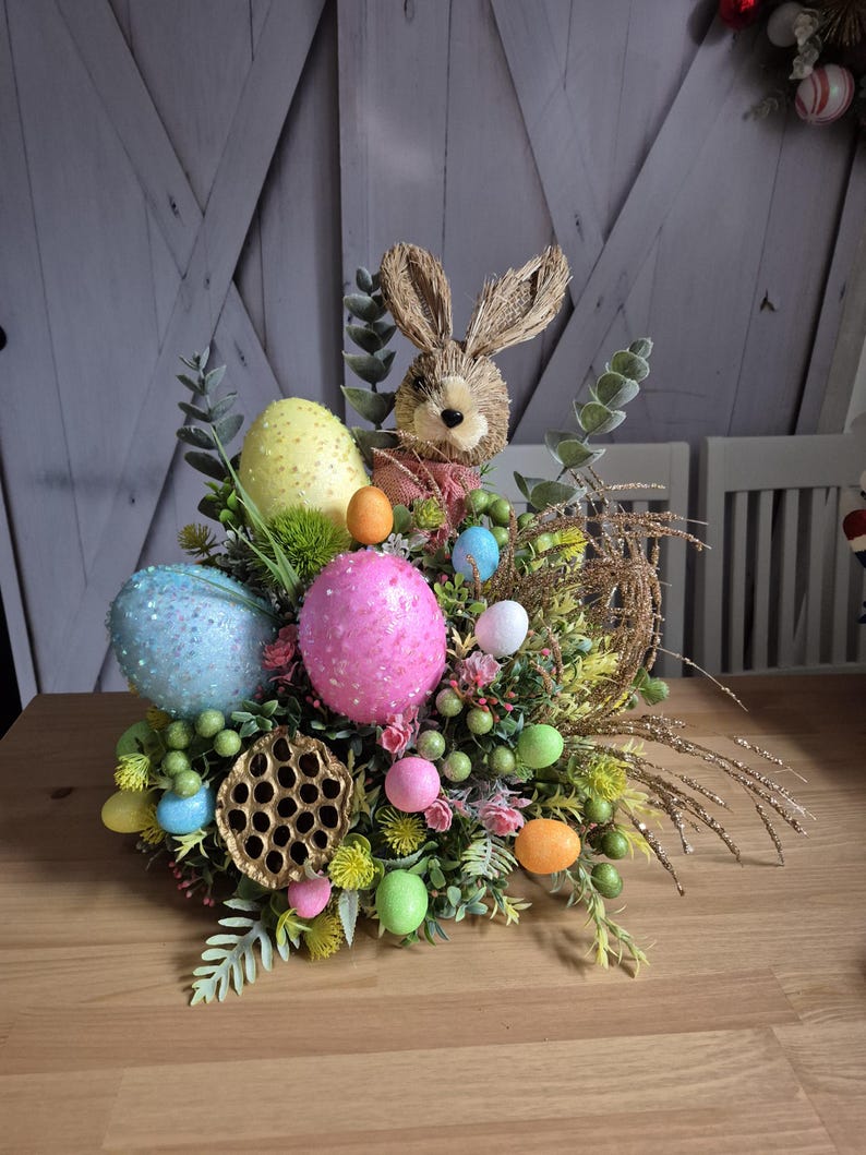 May include: An Easter centrepiece with a straw bunny, colourful eggs, and greenery. The arrangement includes large speckled eggs in blue, yellow, and pink, surrounded by smaller eggs and decorative elements. The centrepiece is set on a wooden table.
