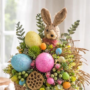 May include: An Easter basket arrangement with a straw bunny, colourful eggs, and greenery. The basket contains blue, pink, and yellow eggs. The arrangement is set in a woven basket, creating a festive spring display.