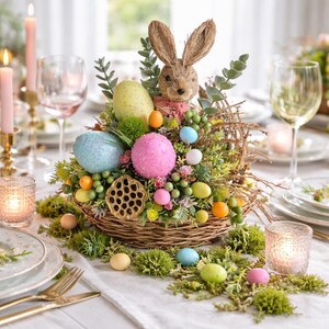 May include: Easter table centrepiece in a woven basket, featuring a straw bunny, colourful eggs, and greenery. The arrangement includes pink, blue, and yellow eggs, with additional decorative elements. The centrepiece is set on a white tablecloth with candles and glassware.
