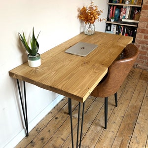 May include: A rustic wooden desk with black hairpin legs. The desk has a natural wood finish and is topped with a laptop computer. A potted plant and a vase of dried flowers are on the desk.