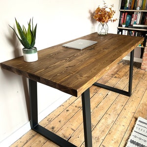 May include: A rustic wooden desk with black metal legs. The desk has a natural wood finish and is shown with a laptop computer on top. The desk is in a room with wood floors and a bookshelf in the background.