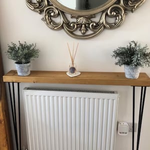 Może przedstawiać: Drewniany stolik konsolowy z czarnymi metalowymi nóżkami w kształcie szpilek do włosów. Stolik jest ozdobiony dwoma doniczkami z roślinami i dyfuzorem z trzciną. Stolik stoi przed białym grzejnikiem.