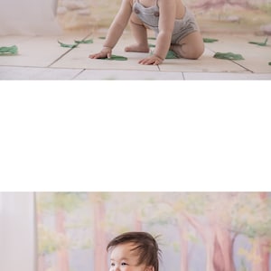 May include: A baby in a gray onesie with brown buttons sits on a white floor with green leaves scattered around. The baby is smiling and looking to the right. The background is a painted mural of a forest with pink, green, and blue colors.
