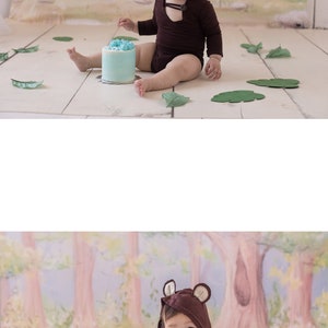May include: A baby wearing a brown bear costume sits on a white floor with a blue and white cake in front of them. The cake has a white topper with the text "Brandie & Co."