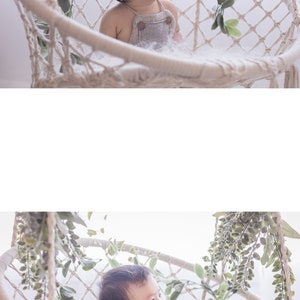 May include: A baby wearing a gray jumpsuit sits in a white macrame swing chair with green leafy vines hanging around the chair.